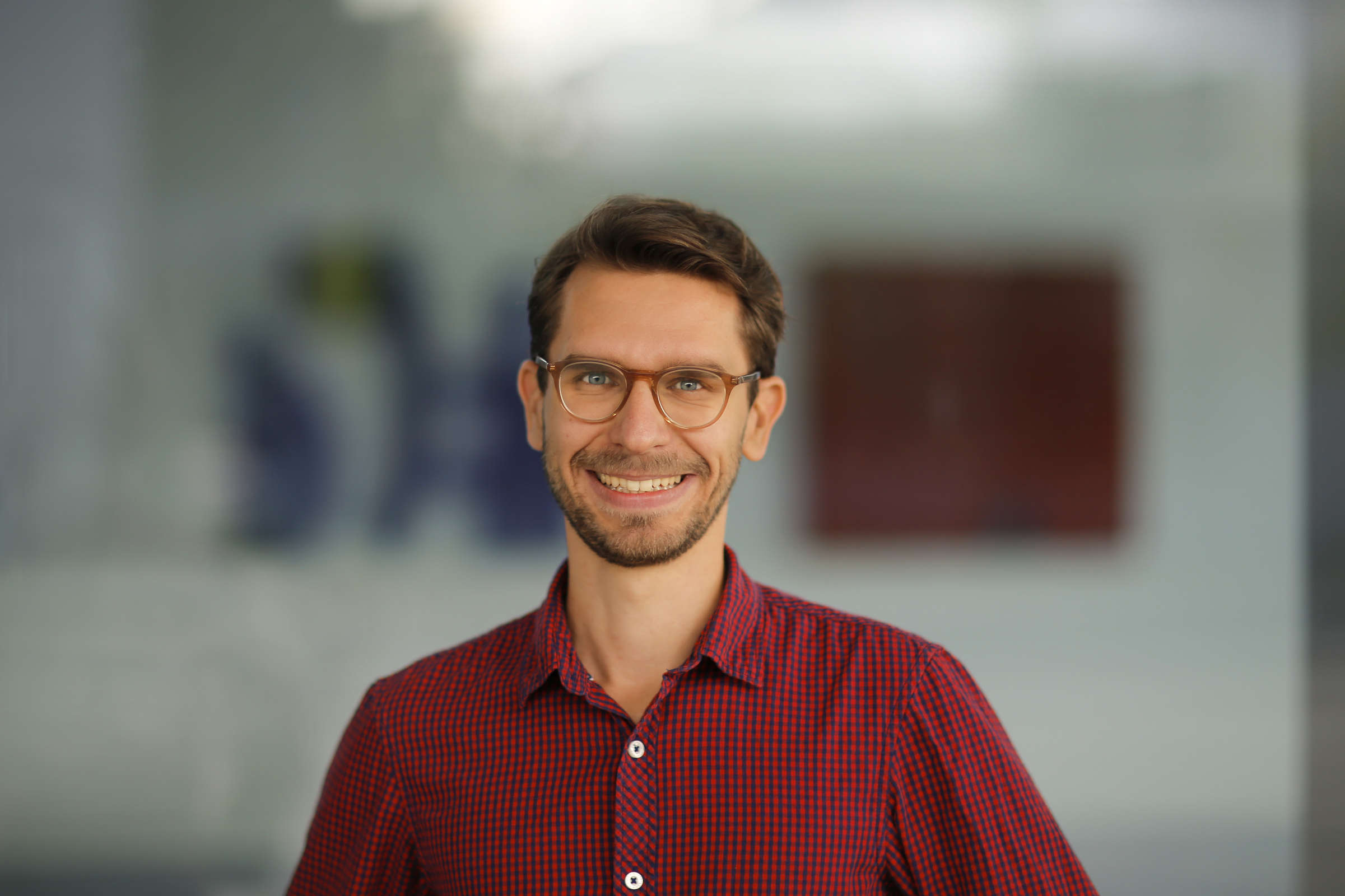 A young white man with short brown hair, glasses and a red shirt smiles into the camera.